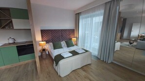 Quarto de hotel moderno com cama, luminárias laterais, cozinha pequena e uma grande janela com cortinas.