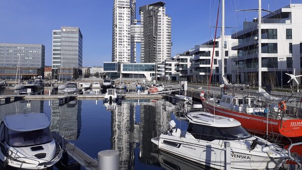 Uma marina com barcos atracados, rodeada por edifícios modernos e um céu azul límpido.