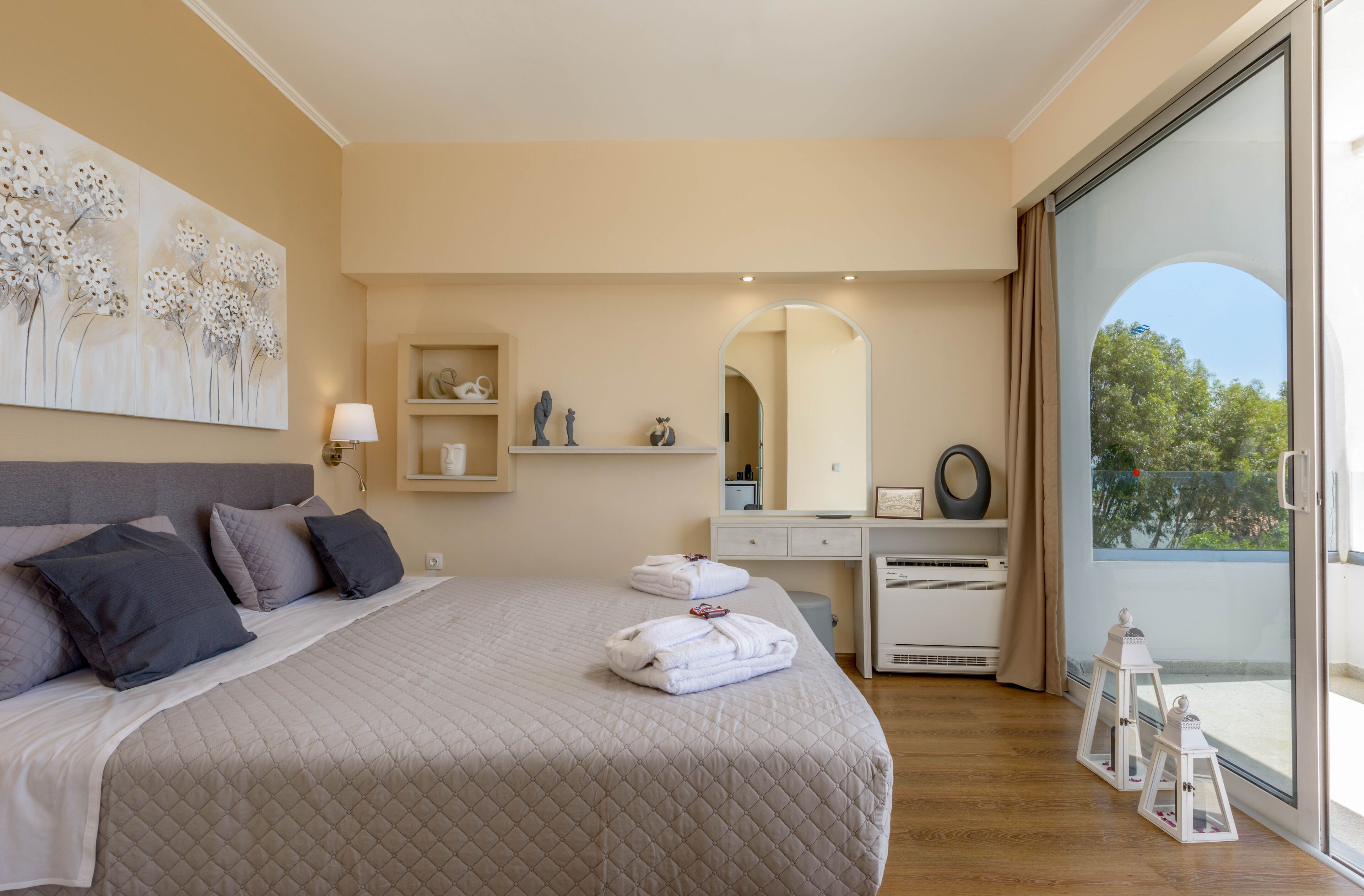 Ein Schlafzimmer mit einem großen Bett, einem Nachttisch, einer Kommode, einem Spiegel und einem Fenster mit Blick auf Bäume.