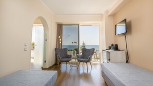 A hotel room with two single beds, a small table with chairs, a TV mounted on the wall, and a balcony with a view of the sea. - Olympos Beach (Rhodes)