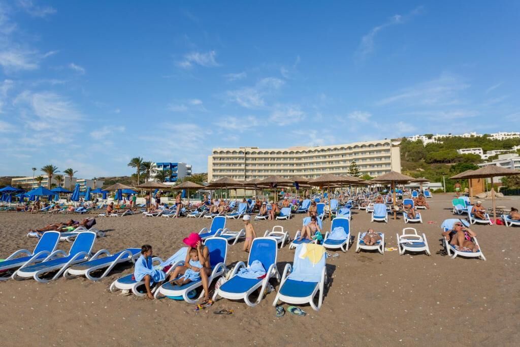 A beach with rows of sun loungers and umbrellas, a large hotel building in the background, and palm trees.