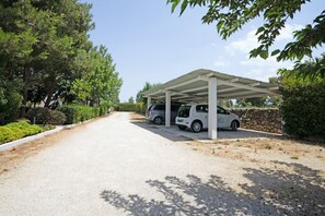 Un espace de stationnement couvert où deux voitures sont garées sous une tonnelle blanche, le tout entouré de verdure et bordé d’un chemin de gravier.