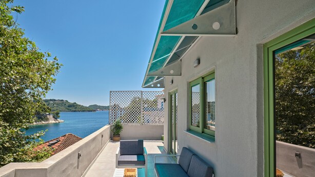 Un balcón con mobiliario exterior, vistas al mar y un edificio con ventanas verdes.
