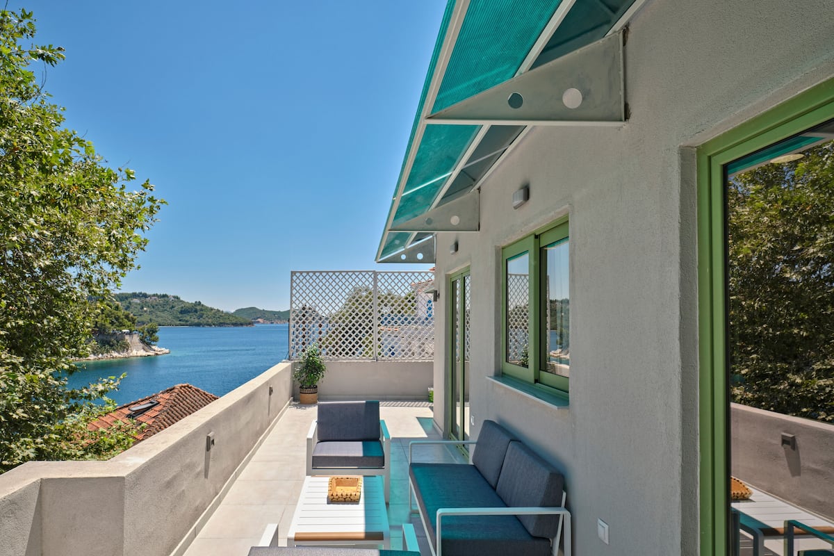 A balcony with outdoor seating, a view of the sea, and a building with green windows.
