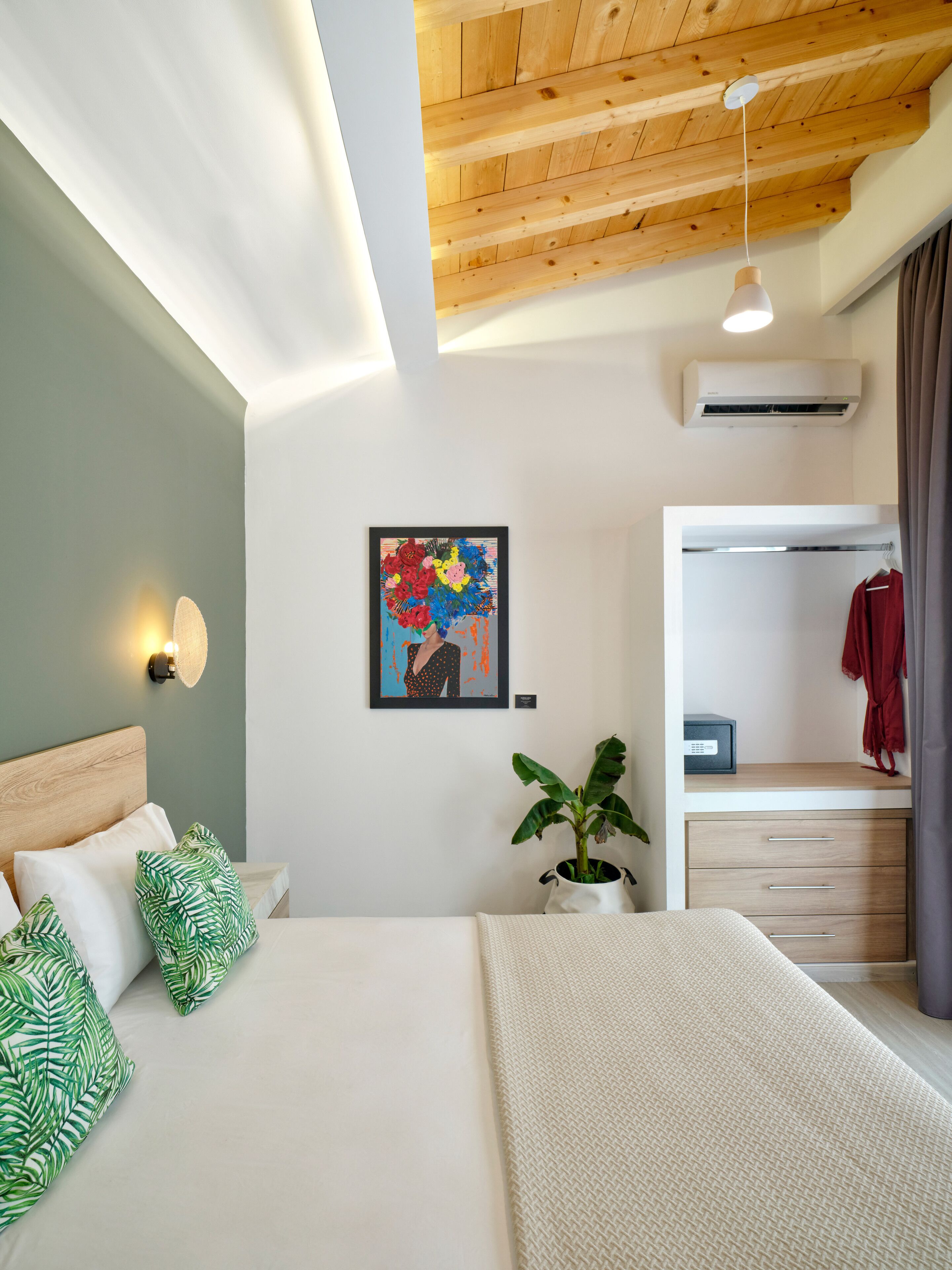 A modern bedroom with a wooden ceiling, a bed with green and white pillows, a potted plant, and a colorful artwork on the wall.