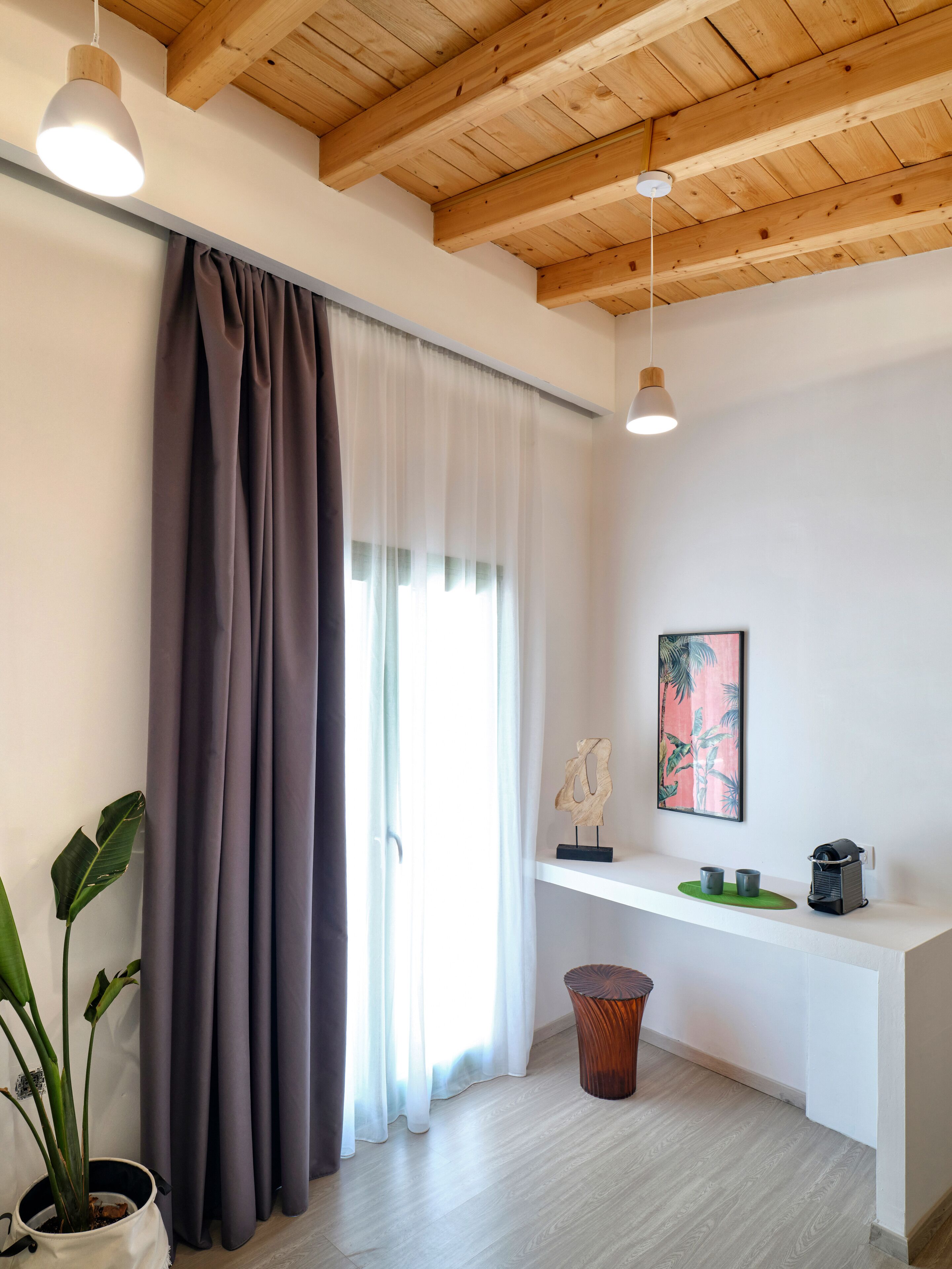 A modern room with a wooden ceiling, a large window with curtains, a plant, a small table with cups, and a wall-mounted shelf with decorative items.