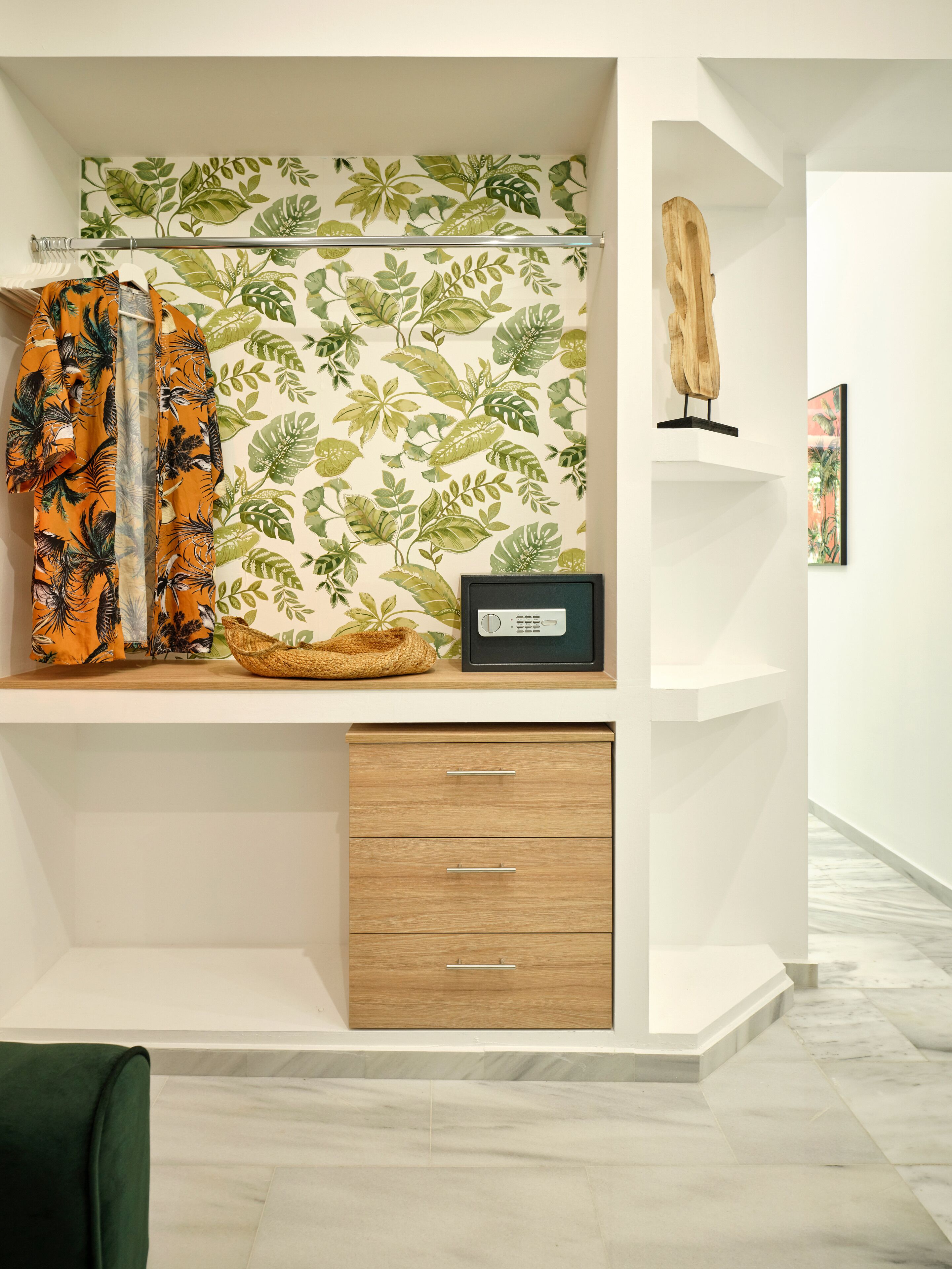 Une chambre moderne avec un mur blanc, une étagère en bois sur laquelle se trouve une radio, un papier peint à motifs de feuilles tropicales et un fauteuil vert.