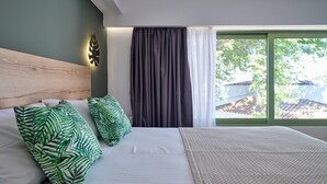 Una cama bien hecha con ropa blanca y almohadas con estampado de hojas verdes, un cabecero de madera y vista a un árbol a través de una ventana con cortinas oscuras.