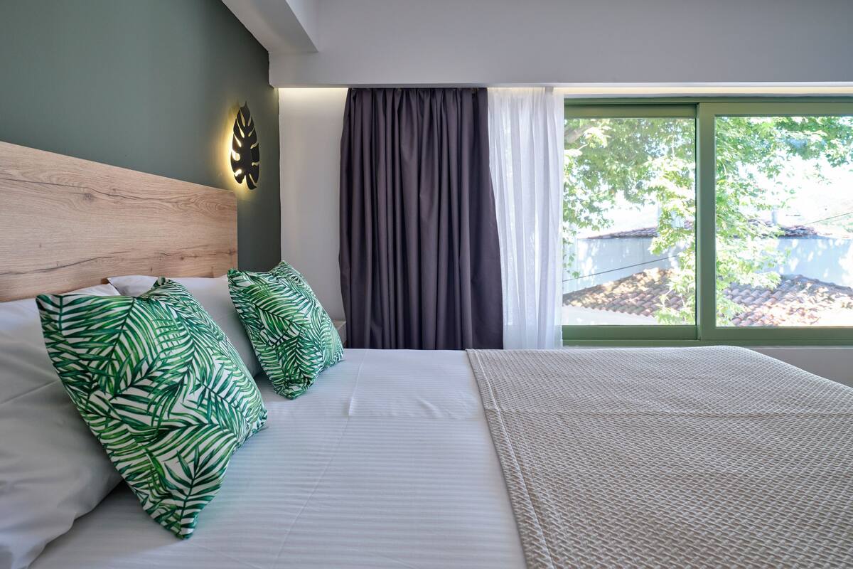 A neatly made bed with white bedding and green leaf-patterned pillows, a wooden headboard, and a view of a tree through a window with dark curtains.