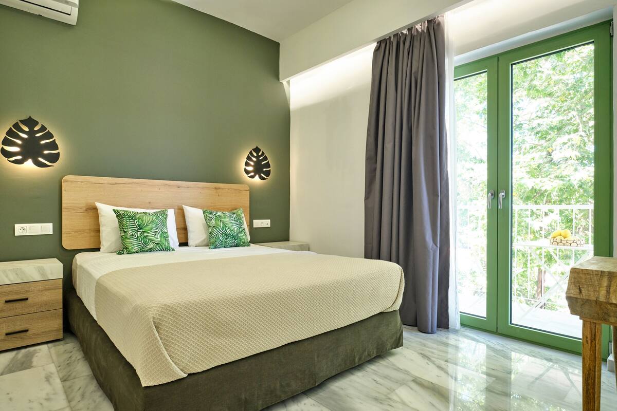 A modern bedroom with a large bed, green walls, and a view of greenery outside.