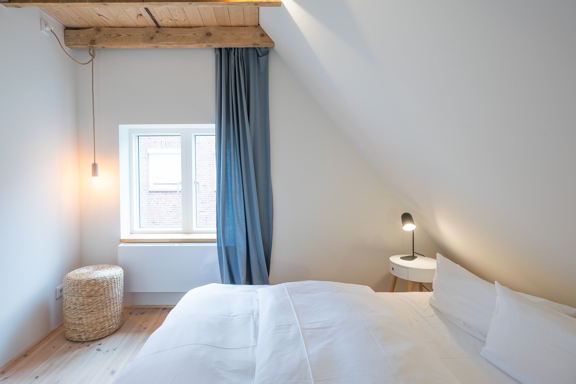 Een slaapkamer met een bed, een nachtkastje met een lamp, een raam met gordijnen en een geweven kruk.