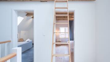 Une échelle en bois dans un couloir, d’où l’on aperçoit une chambre et une salle de bain.