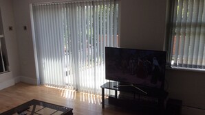 Sala de estar com televisão de tela plana em um suporte, mesa de centro de vidro e porta de correr com persianas verticais.