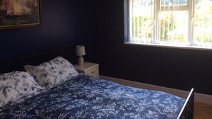 Um quarto com cama, mesa de cabeceira, luminária, janela com vista para vegetação e um quadro na parede.