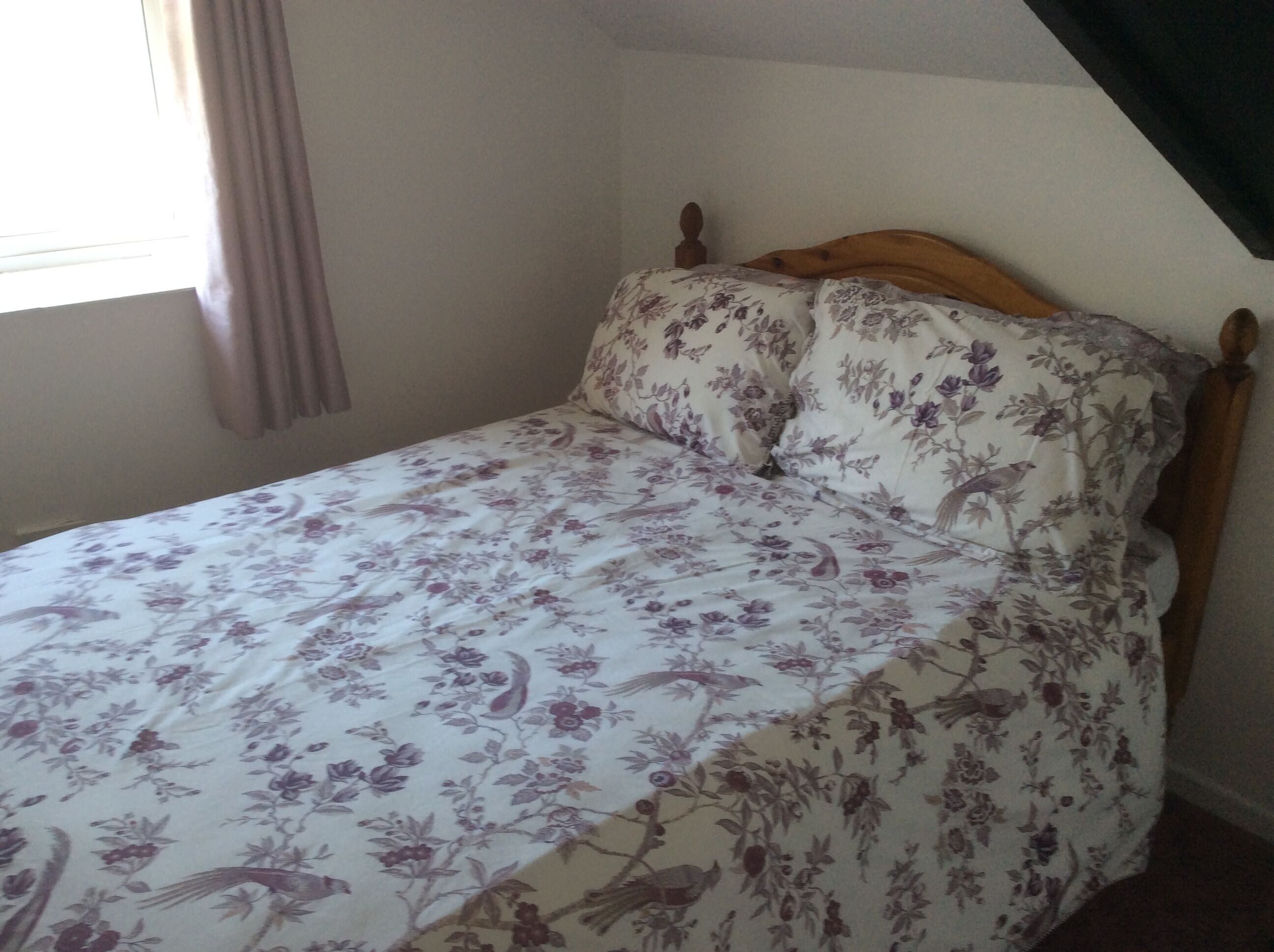 A single bed with a floral bedspread and wooden headboard in a room with a plain wall and a window with curtains.