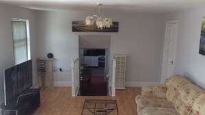 Sala de estar com televisão de tela plana, sofá, mesa de centro e cozinha visível através de uma porta aberta.