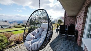 Una silla colgante con asiento y respaldo acolchados, un muro de ladrillo y vistas a un paisaje verde con colinas al fondo.