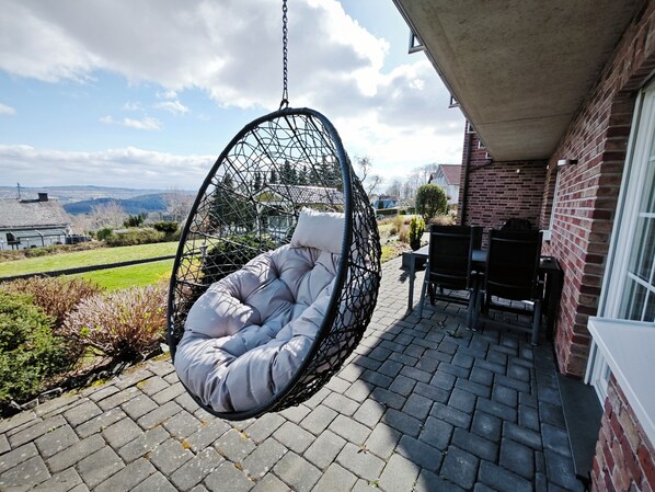 A hanging chair with a cushioned seat and back, a brick wall, and a view of a green landscape with distant hills.