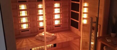 A room with a wooden floor, a bed, and a glass partition. There are windows with blinds and a shelf with towels.