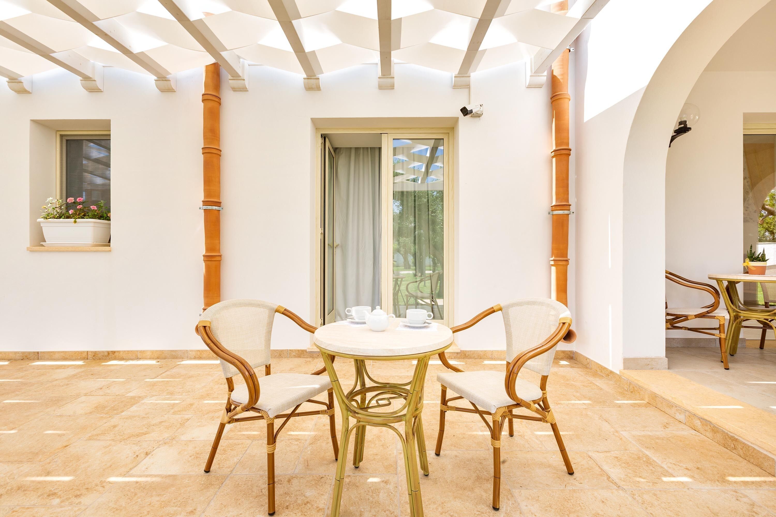 A sunlit patio with a round table and four wicker chairs, a window with a potted plant, and a door with a curtain.