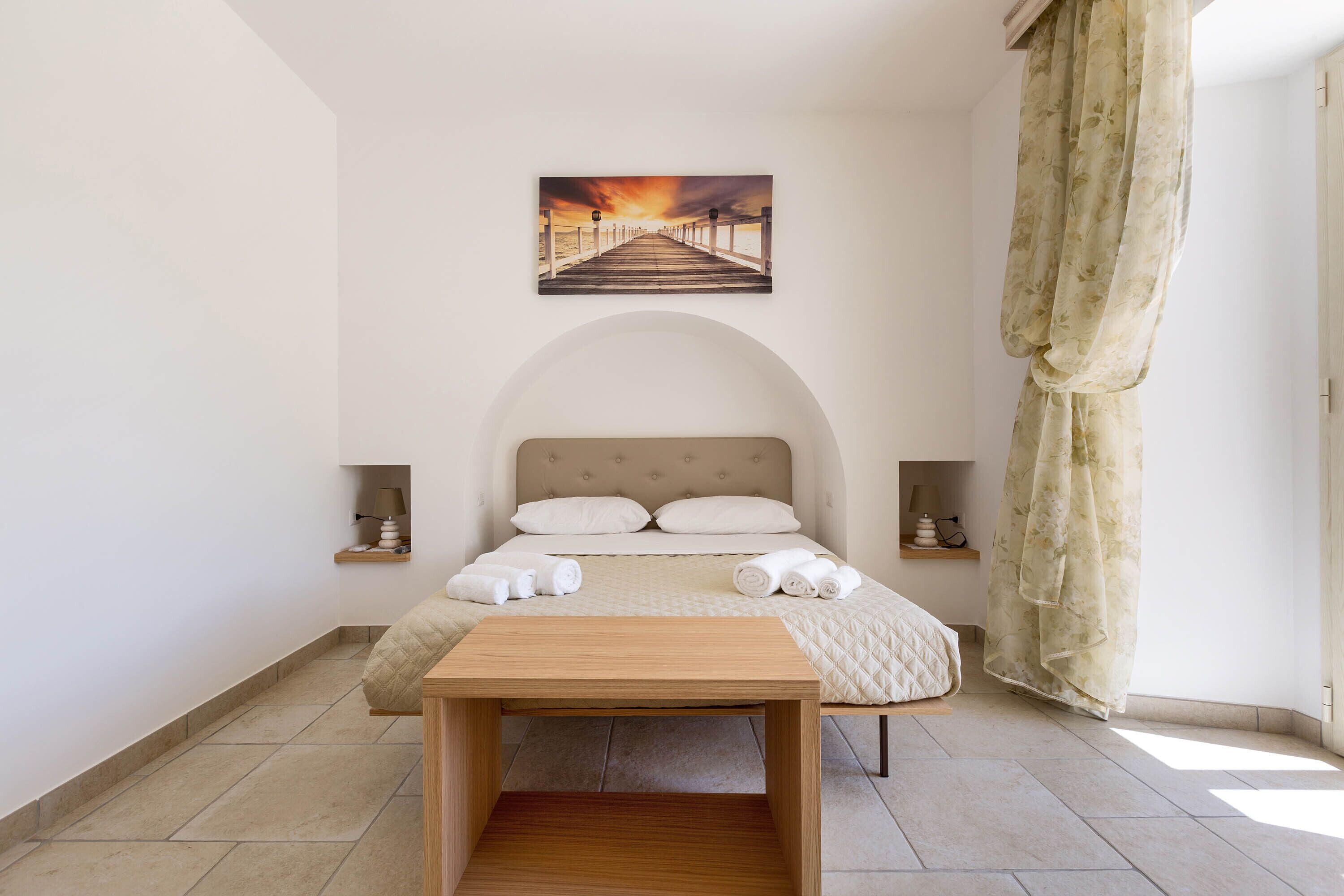 Une chambre à coucher avec un lit, une table en bois, un tableau représentant une jetée et une fenêtre avec des rideaux.
