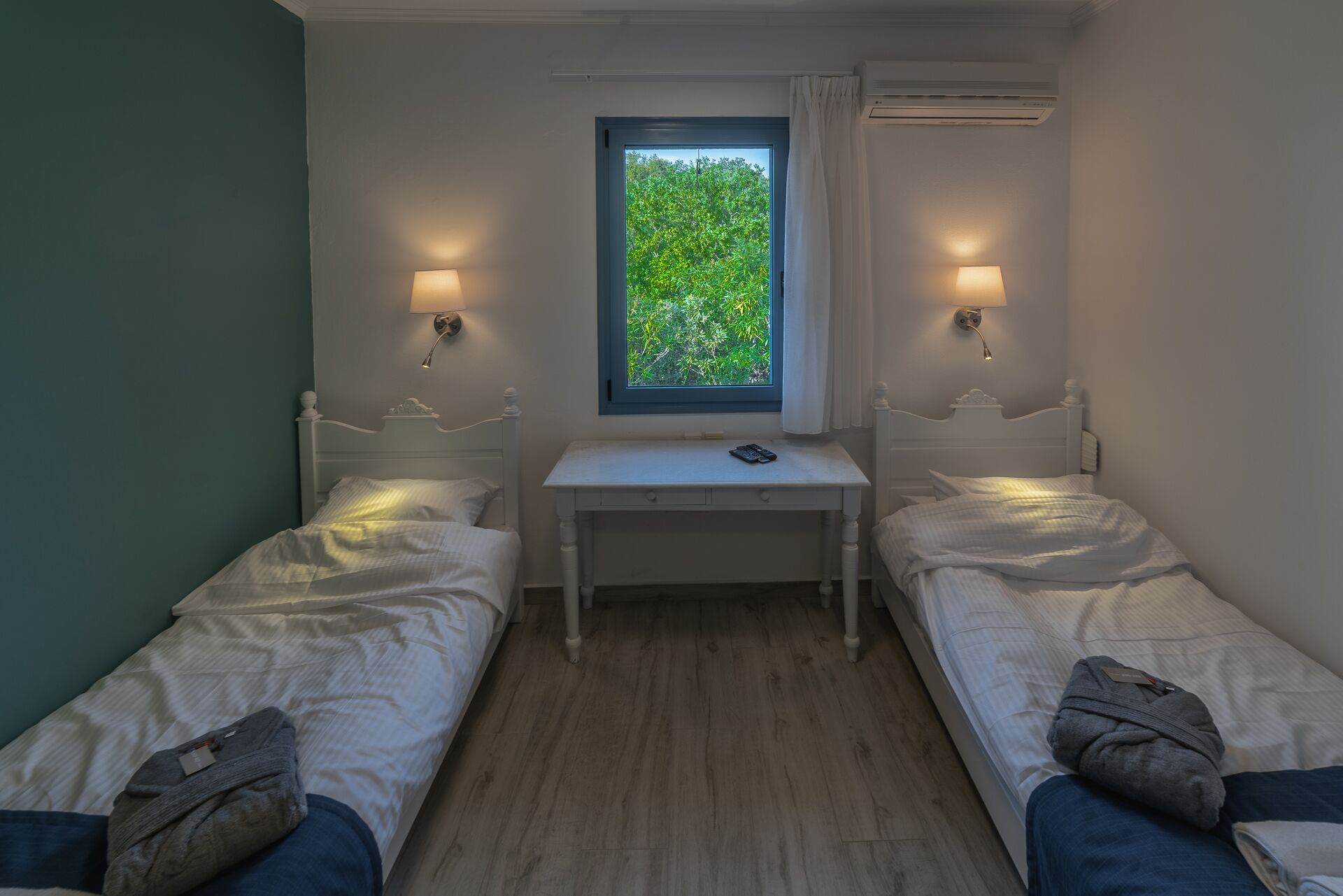 Un dormitorio pequeño y bien iluminado con dos camas individuales, un escritorio y una ventana que da a zonas verdes.