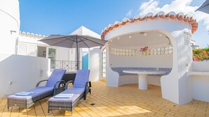Een zonnig buitengebied met twee lounge stoelen en een grote parasol, een decoratieve bank met betegelde basis en een witte muur met een pannendak.