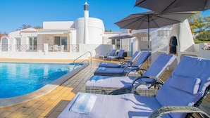 Een zwembadterras met ligstoelen en parasols, een helderblauw zwembad en een gebouw met witte muren en een schoorsteen.