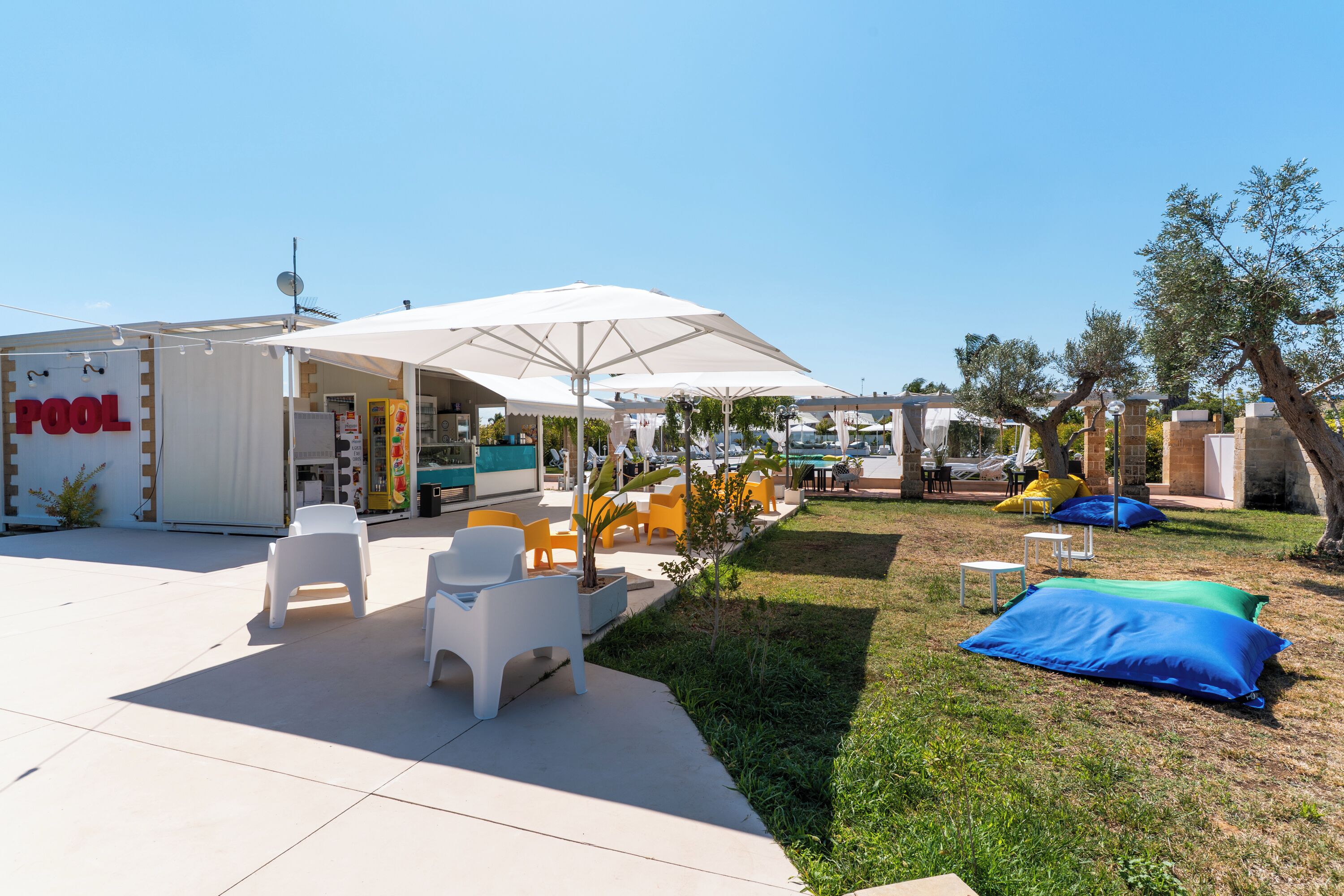 An outdoor pool area with white and yellow chairs, a large umbrella, and a poolside bar.