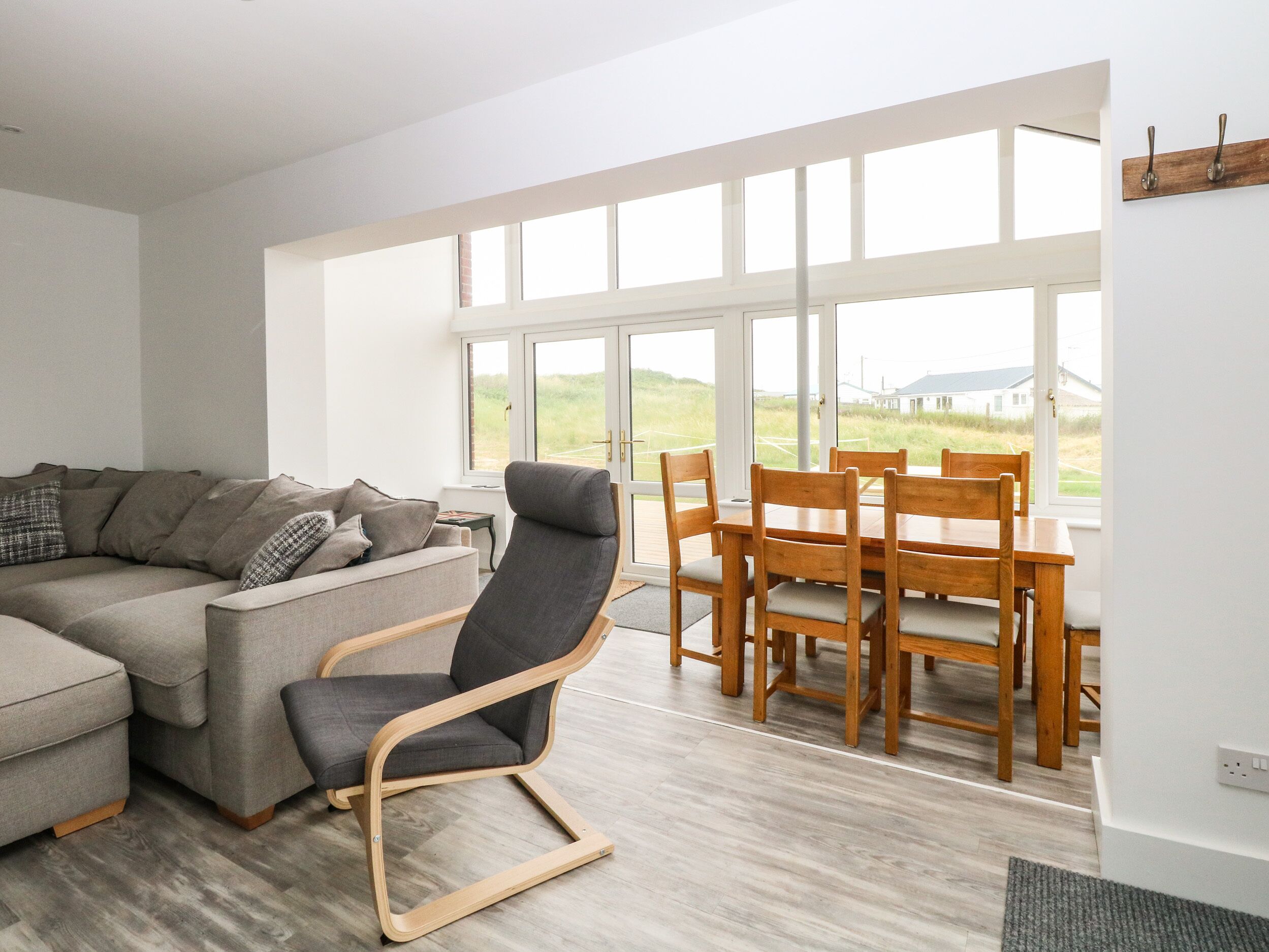 A modern living room with a grey sofa, a wooden dining table with chairs, and a large window offering a view of the outdoors.