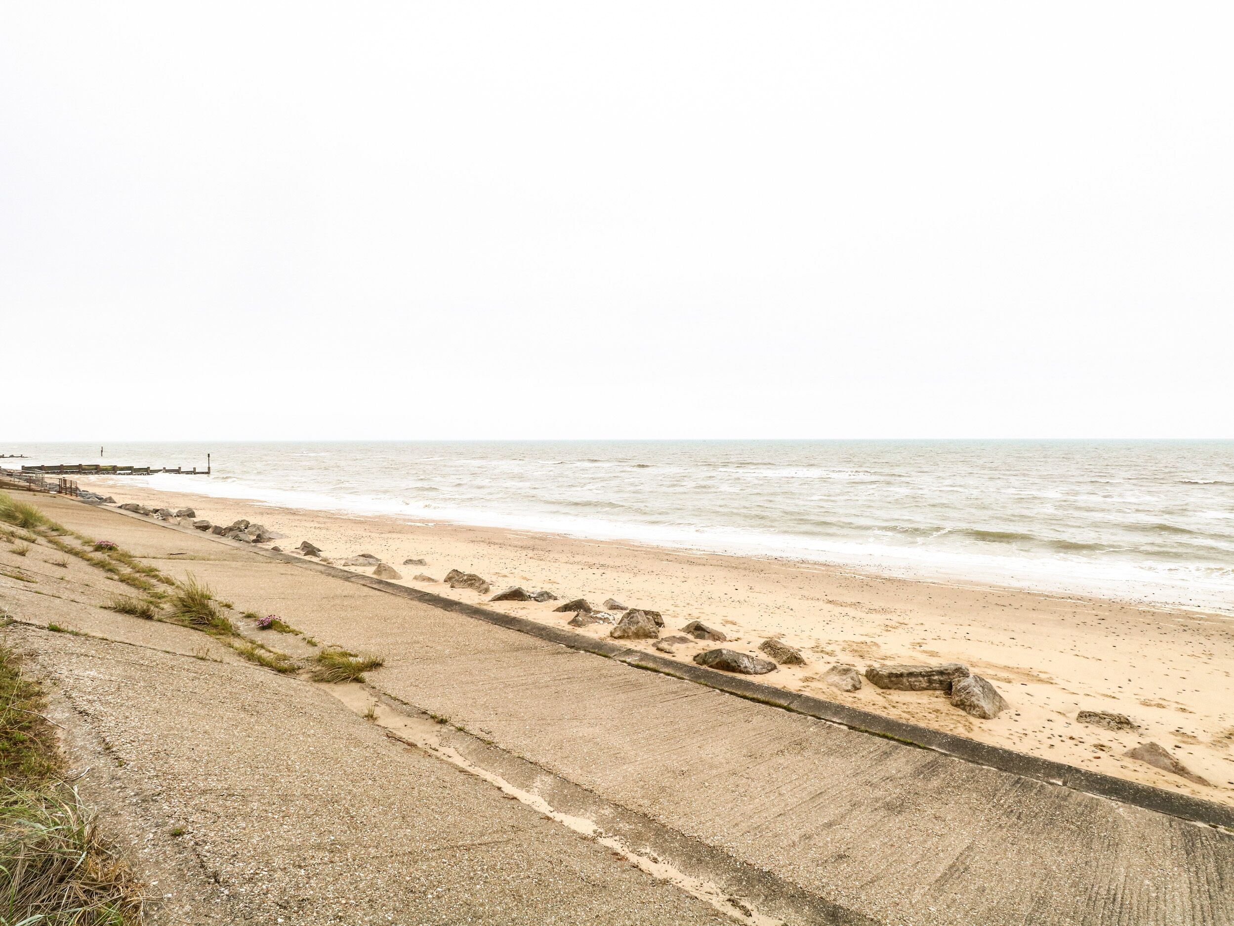 A concrete walkway beside a sandy beach with a rocky shoreline and a pier in the distance.