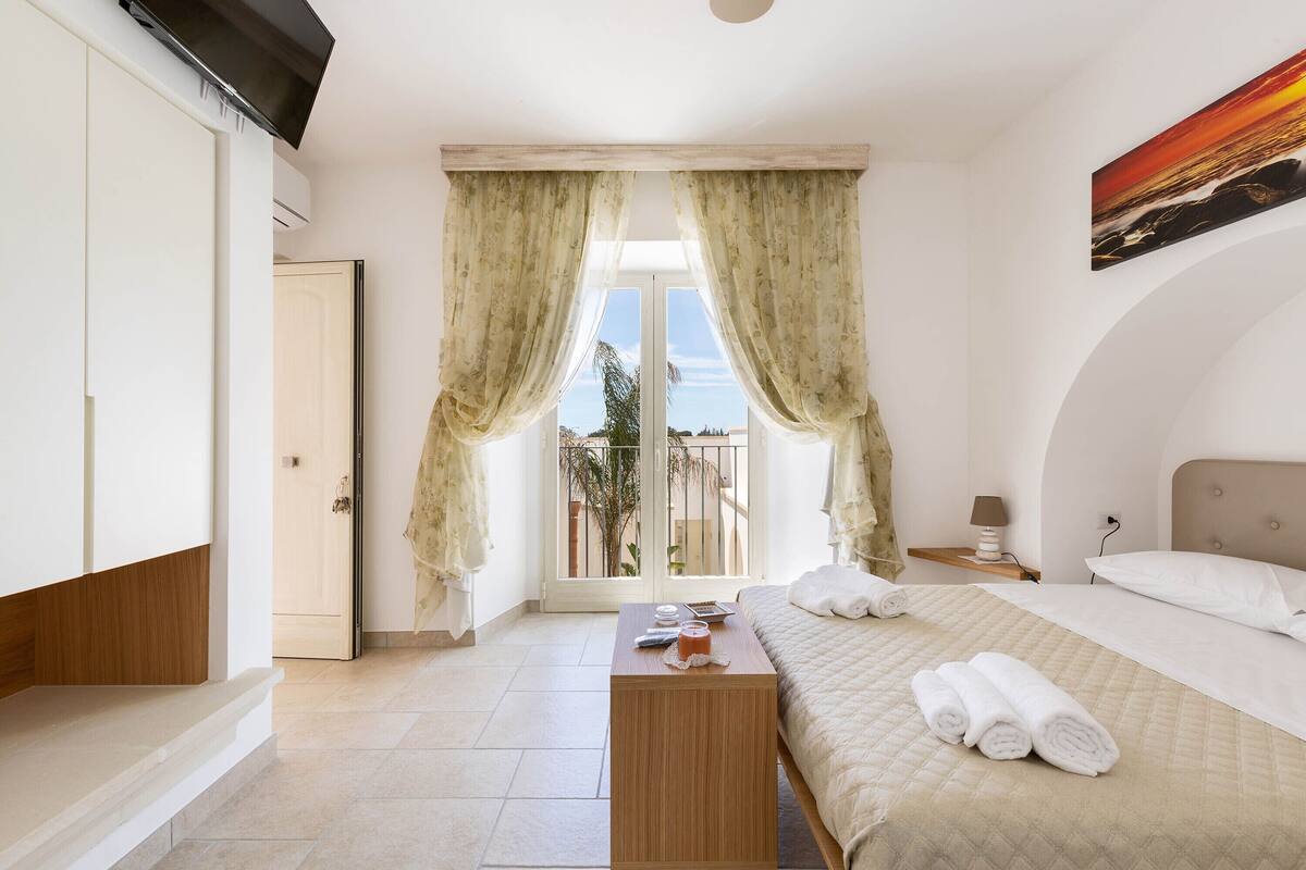 Un dormitorio con cama, una mesita de noche, una ventana con cortinas, un cuadro en la pared y vistas al exterior.