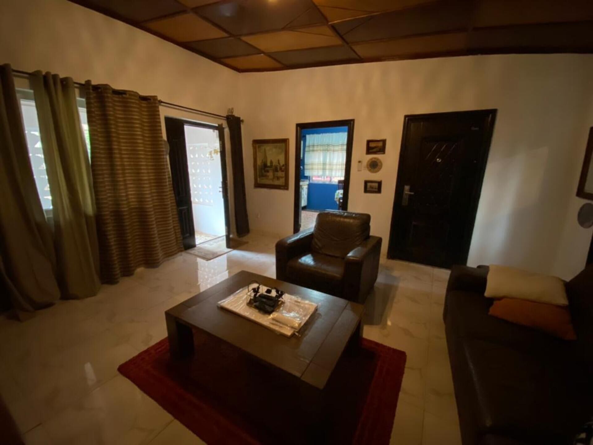 A living room with a brown leather sofa, a wooden coffee table, and a red rug. There is a door and a window with curtains.