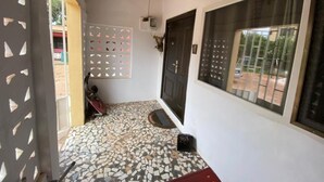 A covered outdoor area with a tiled floor, a dark door, and a window with a metal grille. - Stunning 2-bed House in Madina (Accra)