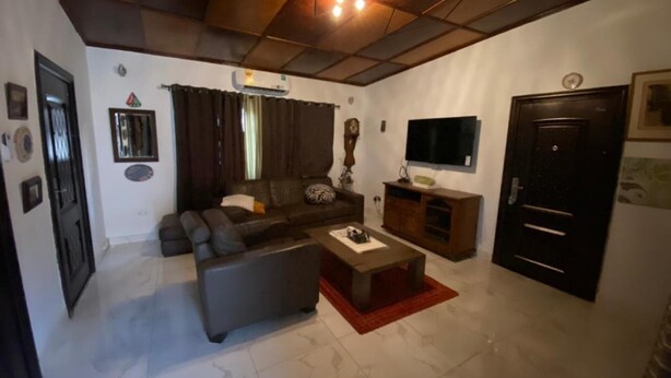 A living room with a sofa, coffee table, and TV. There is a clock and a door on the wall. - Stunning 2-bed House in Madina (Accra)