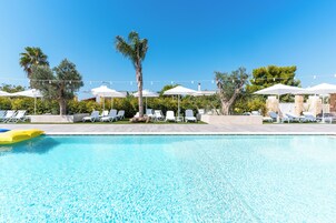 Zona de piscina con sillones de descanso y sombrillas, rodeada de árboles y cielo despejado.
