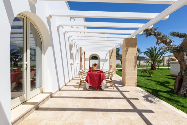 A covered outdoor patio with a table set for dining, surrounded by white columns and a clear blue sky.