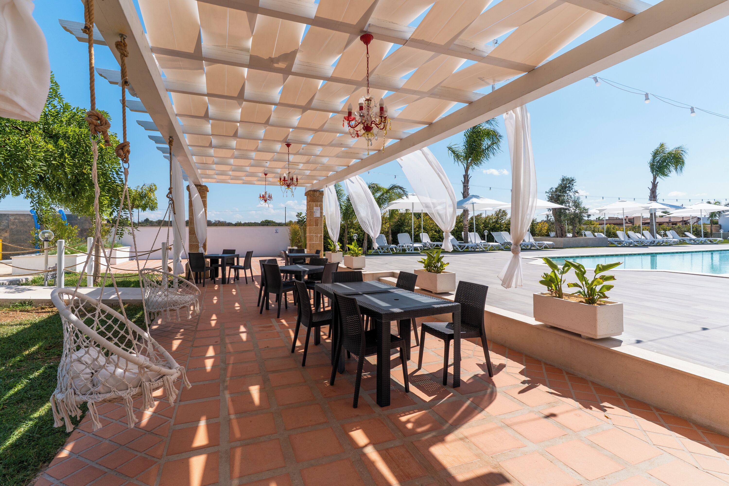 Un espace extérieur aménagé en terrasse avec des tables et des chaises, une piscine en arrière-plan et une pergola décorée de guirlandes lumineuses.