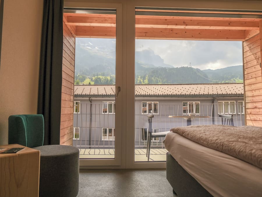 A room with a large window overlooking a balcony with a view of buildings and mountains.