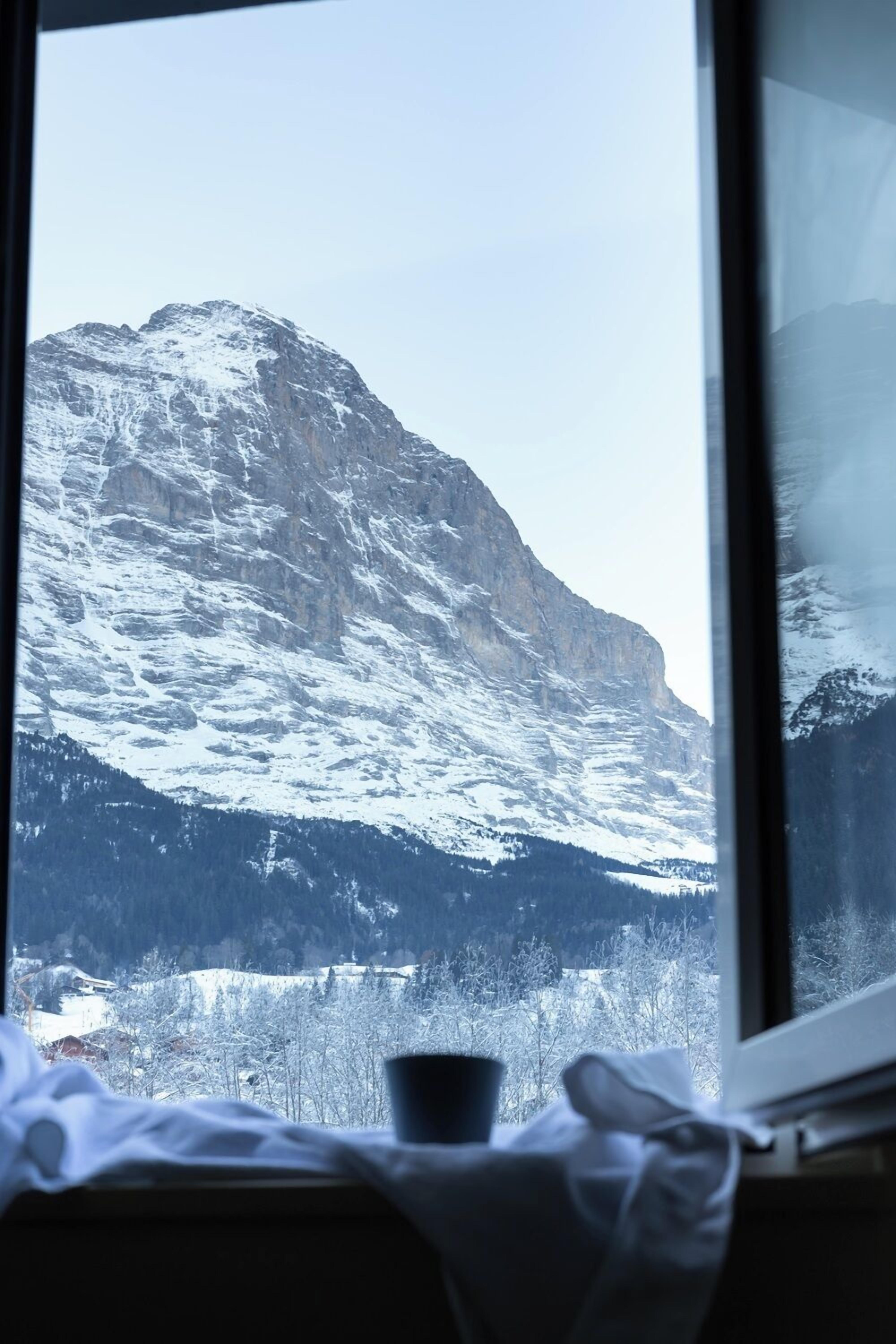 A mountain view through a window with a cup on the windowsill.