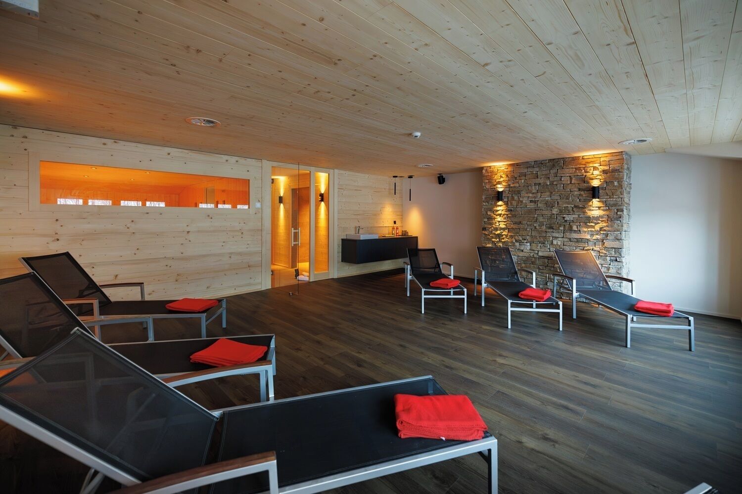 Une salle de spa moderne avec des murs en bois, un mur d’accent en pierre et plusieurs chaises longues, toutes dotées de serviettes rouges.