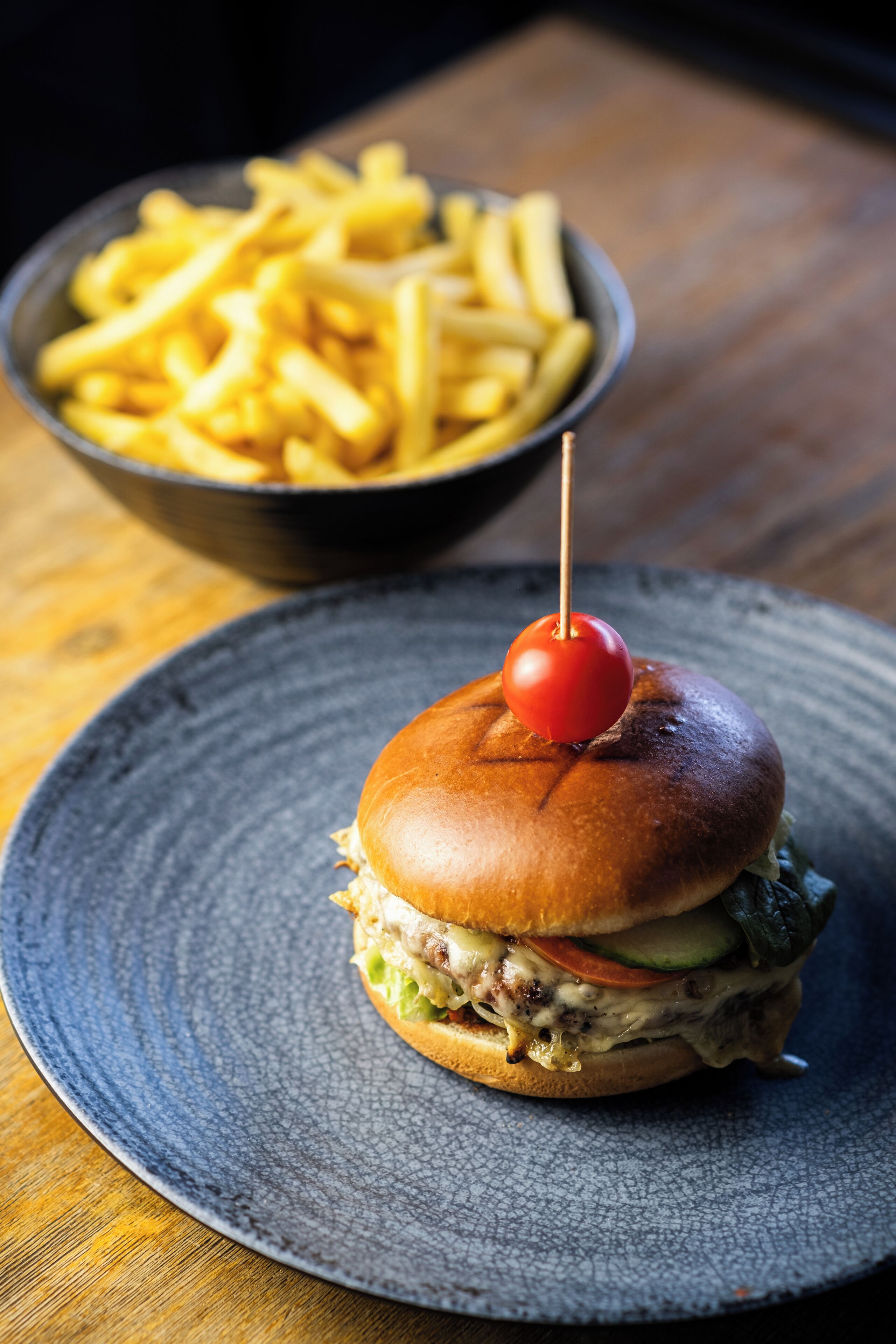 Hamburguesa con cereza encima, servida en un plato con un bol de papas fritas al fondo.
