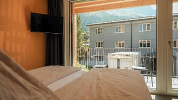 Een slaapkamer met een bed, een aan de muur bevestigde televisie en een balkon met uitzicht op een gebouw en bergen.