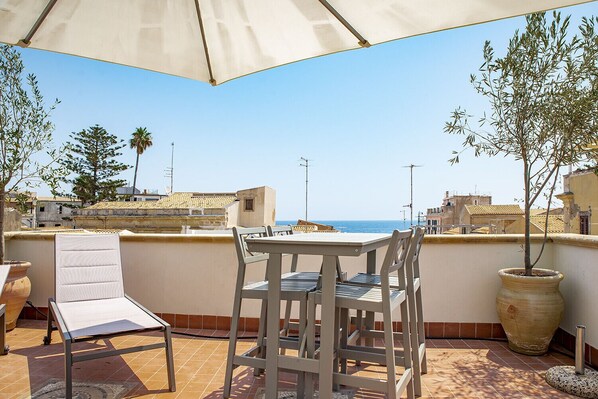 A terrace with a table and chairs, overlooking a coastal view with buildings and a clear sky. - La Terrazza sul Vicolo della Giudecca by Wonderful Italy (Siracusa)