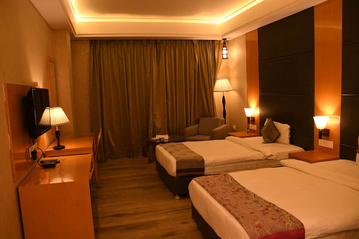 Quarto de hotel com duas camas, uma escrivaninha com luminária, uma cadeira, televisão e uma janela grande com cortinas.