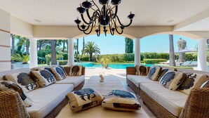 A covered outdoor seating area with wicker furniture, a pool in the background, and a view of the landscape.