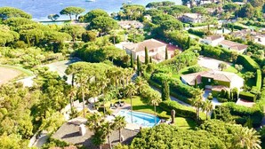 A coastal resort with a swimming pool, lush greenery, and villas with red-tiled roofs.