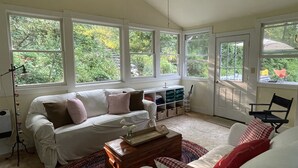 A cozy living room with a white sofa, a wooden coffee table, and a view of greenery through large windows. - Peacock Garden Cottage - Discounted for Winter Month Rental (Pine plains)