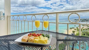 Un balcon avec une table dressée, sur laquelle se trouve une assiette de nourriture, deux verres de jus d’orange, et d’où l’on peut admirer la plage et l’océan.