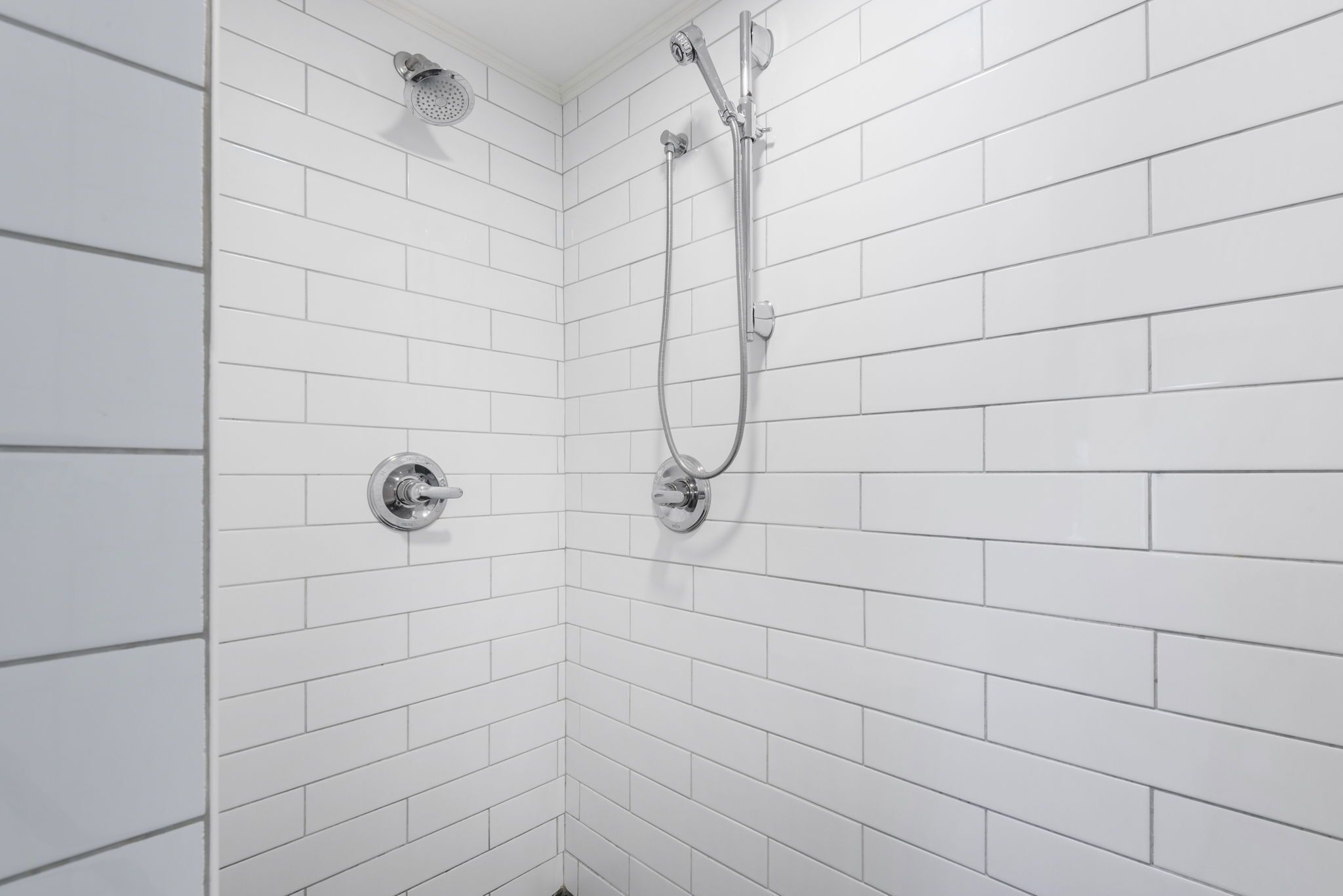 A shower with a handheld showerhead and a wall-mounted faucet in a corner with white tiled walls.